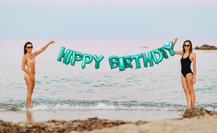 two women holding a green happy birthday balloon sign at the beach