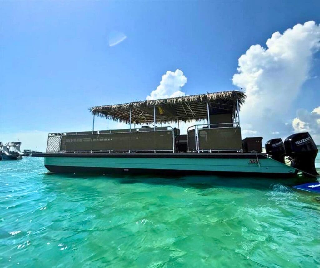 A Crab Island Tiki Boat with a thatched roof floats on turquoise water under a sunny sky.