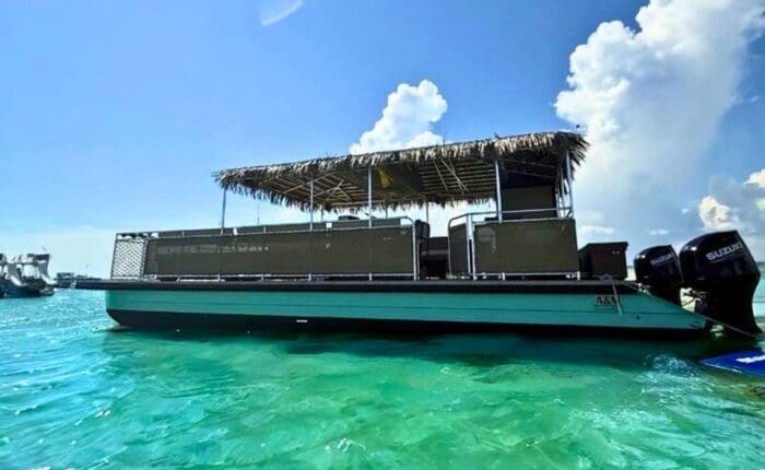 A Crab Island Tiki Boat with a thatched roof floats on turquoise water under a sunny sky.